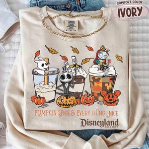 May include: Ivory-colored sweatshirt with a graphic featuring characters and pumpkin spice drinks. The text reads "Pumpkin Spice & Everything Nice Disneyland Resort". The sweatshirt has a crew neck and long sleeves.
