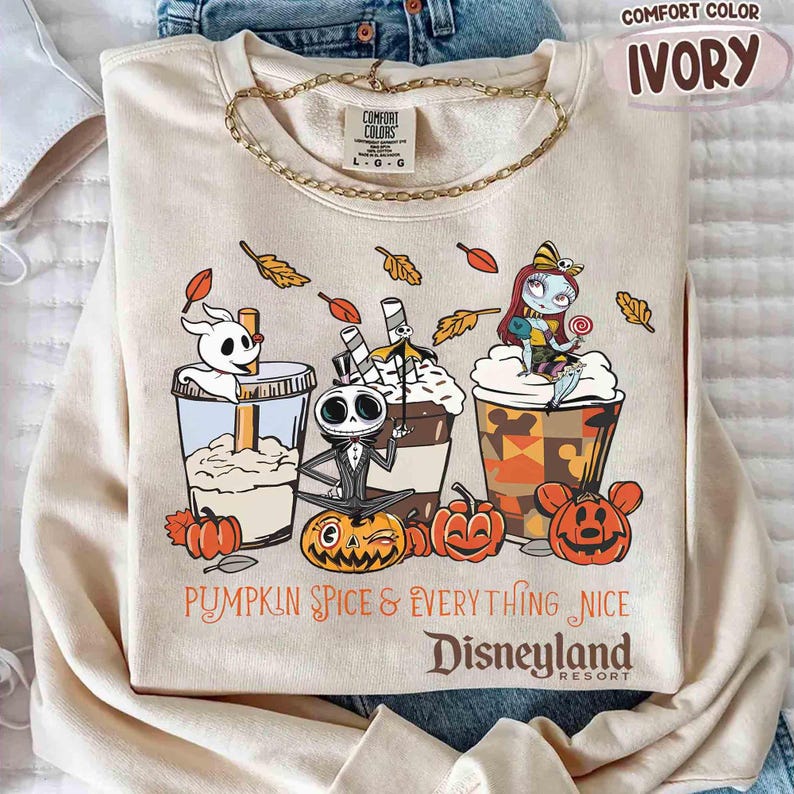 May include: Ivory-colored sweatshirt with a graphic featuring characters and pumpkin spice drinks. The text reads "Pumpkin Spice & Everything Nice Disneyland Resort". The sweatshirt has a crew neck and long sleeves.