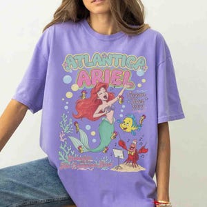 May include: A light purple t-shirt featuring a graphic print of Ariel the mermaid, Flounder, and Sebastian. The text reads Atlantica Ariel Ocean Tour 1991 and Sebastian with the Hot Crustacean Band, surrounded by bubbles, musical notes, and coral.