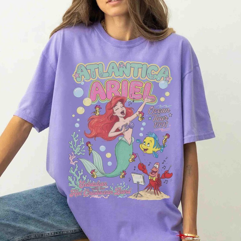 May include: A light purple t-shirt featuring a graphic print of Ariel the mermaid, Flounder, and Sebastian. The text reads Atlantica Ariel Ocean Tour 1991 and Sebastian with the Hot Crustacean Band, surrounded by bubbles, musical notes, and coral.