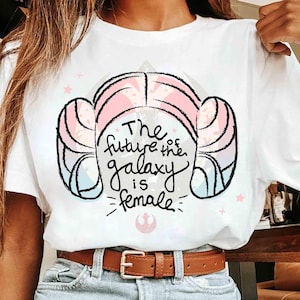 Può includere: T-shirt bianca con una grafica di un'acconciatura con accenti rosa e blu, e la scritta "The future of the galaxy is female." Il design include stelle e un simbolo. La maglietta è indossata con una cintura marrone.