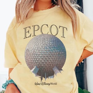 May include: A pale yellow short-sleeve t-shirt featuring a large graphic of the Epcot Spaceship Earth geodesic sphere. The word EPCOT is printed in a black serif font above the sphere, and the text Walt Disney World is printed below it.
