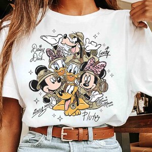 May include: White t-shirt with a colourful graphic of Disney characters Mickey Mouse, Minnie Mouse, Donald Duck, Daisy Duck, Goofy, and Pluto in safari outfits. The names of the characters are written in cursive. The shirt has a brown belt.