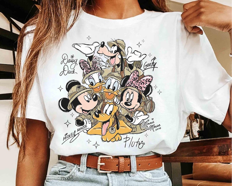 May include: White t-shirt with a colourful graphic of Disney characters Mickey Mouse, Minnie Mouse, Donald Duck, Daisy Duck, Goofy, and Pluto in safari outfits. The names of the characters are written in cursive. The shirt has a brown belt.