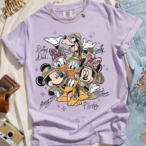 May include: A lilac t-shirt with a graphic of Disney characters Mickey Mouse, Minnie Mouse, Donald Duck, Daisy Duck, Goofy, and Pluto in safari attire. The shirt has the text "Mickey Mouse" and "Minnie Mouse 2020".