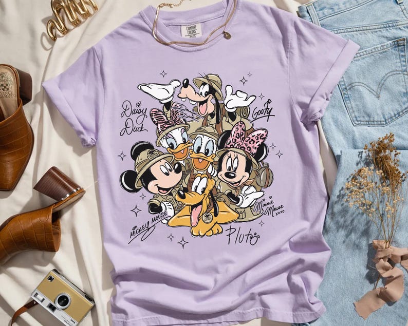 May include: A lilac t-shirt with a graphic of Disney characters Mickey Mouse, Minnie Mouse, Donald Duck, Daisy Duck, Goofy, and Pluto in safari attire. The shirt has the text "Mickey Mouse" and "Minnie Mouse 2020".