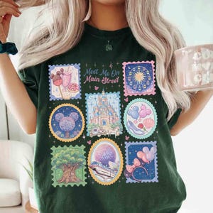 Peut inclure: T-shirt vert fonc&eacute; avec des motifs de timbres-poste color&eacute;s repr&eacute;sentant des images de parc &agrave; th&egrave;me. Les timbres comprennent un ch&acirc;teau, une grande roue et des ballons. Le texte sur le t-shirt se lit "Meet Me On Main Street."