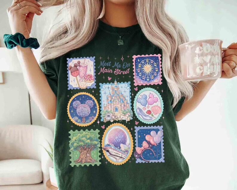Peut inclure: T-shirt vert fonc&eacute; avec des motifs de timbres-poste color&eacute;s repr&eacute;sentant des images de parc &agrave; th&egrave;me. Les timbres comprennent un ch&acirc;teau, une grande roue et des ballons. Le texte sur le t-shirt se lit "Meet Me On Main Street."