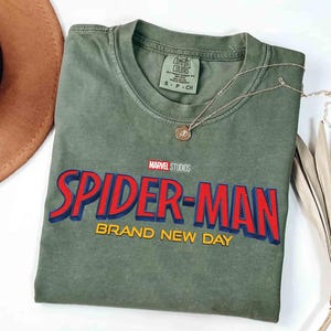 May include: An olive green t-shirt featuring the Marvel Studios Spider-Man logo in red and blue with yellow accents. The words "BRAND NEW DAY" are below the logo. A gold necklace with a pendant is draped across the shirt.