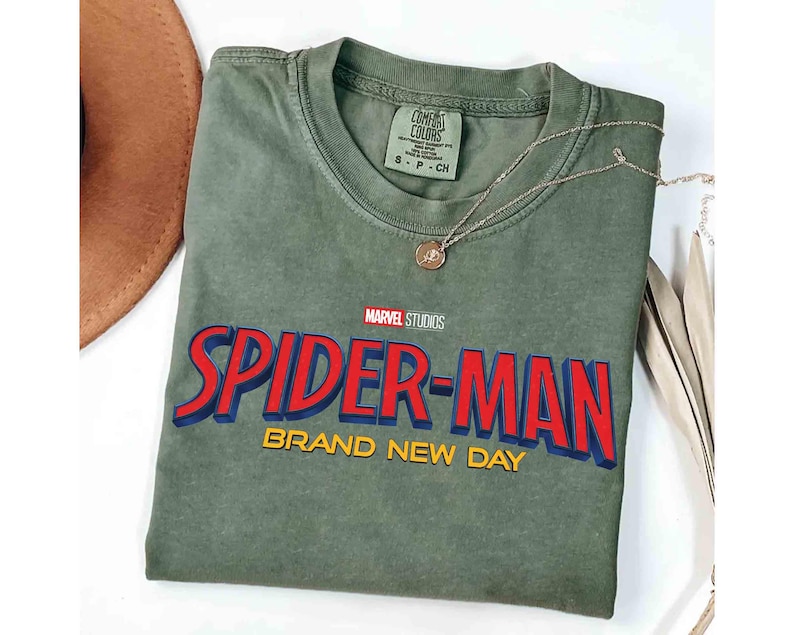 May include: An olive green t-shirt featuring the Marvel Studios Spider-Man logo in red and blue with yellow accents. The words "BRAND NEW DAY" are below the logo. A gold necklace with a pendant is draped across the shirt.
