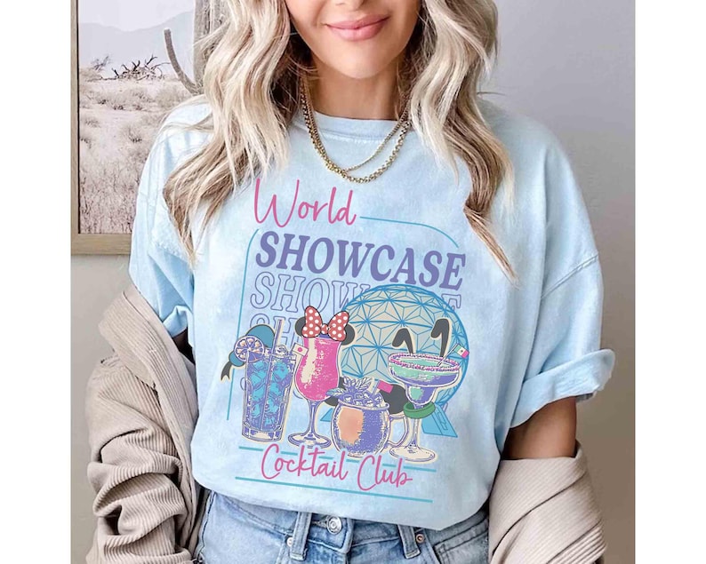 以下が含まれることがあります： 「World Showcase Show Cocktail Club」の文字とカクテルと地球のグラフィックが描かれた水色のTシャツ。袖はロールアップされており、ゆったりとしたフィット感です。
