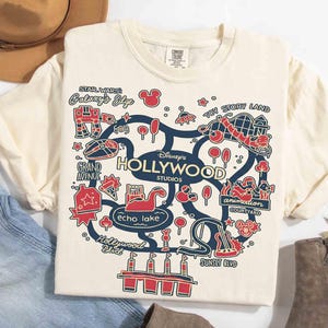May include: Cream-colored t-shirt featuring a map of Disney's Hollywood Studios with red and blue illustrations. The map includes text such as "Star Wars Galaxy's Edge," "Toy Story Land," and "Sunset Blvd." The shirt is folded, with a hat and jeans in the background.