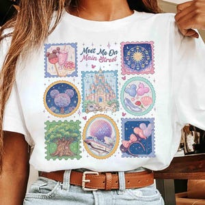 Peut inclure: T-shirt blanc avec un motif de timbres color&eacute;s et le texte "Meet Me On Main Street". Les timbres repr&eacute;sentent diverses ic&ocirc;nes de parc &agrave; th&egrave;me, dont un ch&acirc;teau, une grande roue et des &eacute;l&eacute;ments en forme de Mickey Mouse. Le t-shirt a un col rond classique.