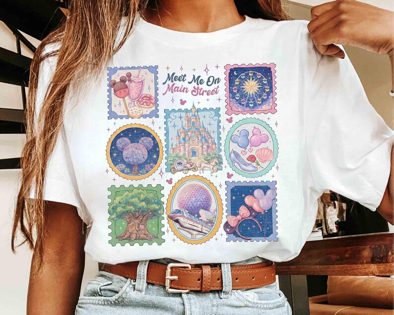 Peut inclure: T-shirt blanc avec un motif de timbres color&eacute;s et le texte "Meet Me On Main Street". Les timbres repr&eacute;sentent diverses ic&ocirc;nes de parc &agrave; th&egrave;me, dont un ch&acirc;teau, une grande roue et des &eacute;l&eacute;ments en forme de Mickey Mouse. Le t-shirt a un col rond classique.