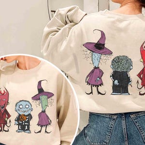 May include: Beige sweatshirt featuring a colorful cartoon illustration of three characters. The characters include a witch in a purple dress and hat, a character in a black suit, and a red devil. The design is on the front and back of the sweatshirt.