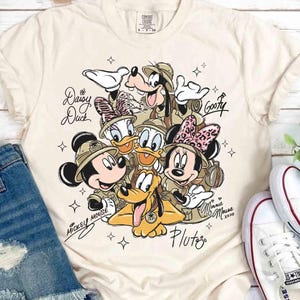 May include: A cream-coloured t-shirt with a graphic of Disney characters Mickey Mouse, Minnie Mouse, Donald Duck, Daisy Duck, Goofy, and Pluto. The characters are wearing safari hats and are surrounded by stars. The shirt has the text "Daisy Duck", "Goofy", "Minnie xoxo", "Pluto", and "Mickey Mouse".