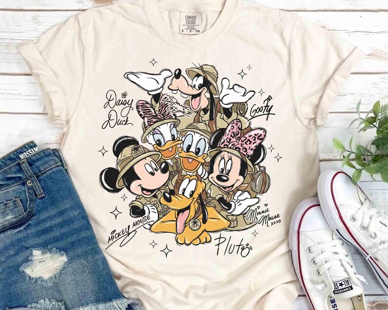 May include: A cream-coloured t-shirt with a graphic of Disney characters Mickey Mouse, Minnie Mouse, Donald Duck, Daisy Duck, Goofy, and Pluto. The characters are wearing safari hats and are surrounded by stars. The shirt has the text "Daisy Duck", "Goofy", "Minnie xoxo", "Pluto", and "Mickey Mouse".
