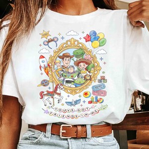 May include: White t-shirt featuring a colorful graphic of characters from a popular animated movie. The design includes a gold frame with characters, a rocket, a carousel horse, and the text "YOU'VE GOT FRIENDS IN ME."