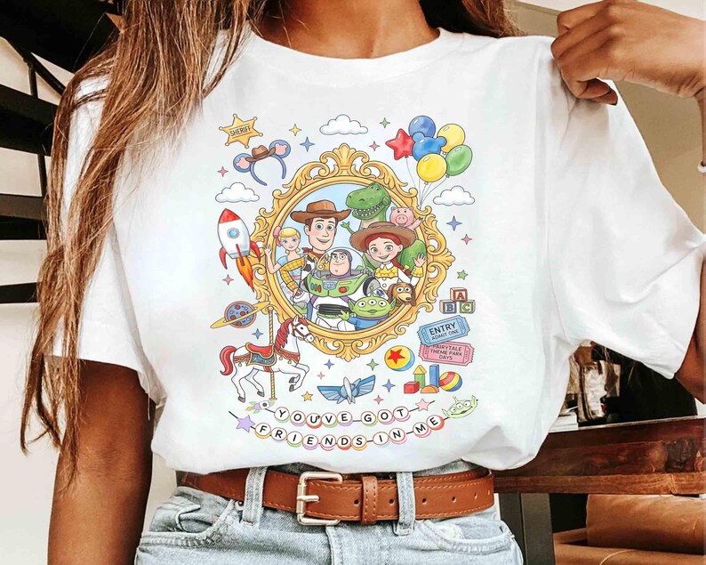 May include: White t-shirt featuring a colorful graphic of characters from a popular animated movie. The design includes a gold frame with characters, a rocket, a carousel horse, and the text "YOU'VE GOT FRIENDS IN ME."