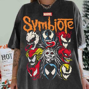 May include: Dark gray t-shirt featuring the Marvel logo and the word "Symbiote" in orange. The shirt displays a collage of colorful, stylized character faces. The design includes red, white, yellow, and blue accents.