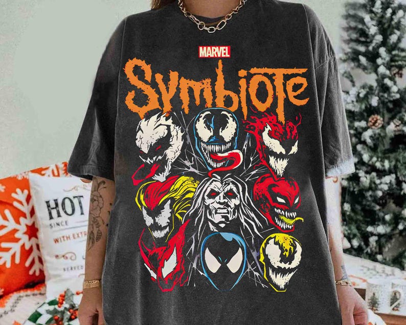 May include: Dark gray t-shirt featuring the Marvel logo and the word "Symbiote" in orange. The shirt displays a collage of colorful, stylized character faces. The design includes red, white, yellow, and blue accents.
