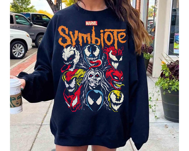 May include: A navy blue sweatshirt featuring the word "Symbiote" in orange, with the Marvel logo above. The sweatshirt displays a collage of colorful character faces. A casual, comfortable garment.