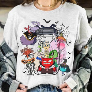 May include: A light gray t-shirt featuring a Halloween-themed graphic. The graphic depicts various cartoon characters inside a glass jar, surrounded by bats, spiderwebs, and spiders. The characters are in various Halloween costumes.
