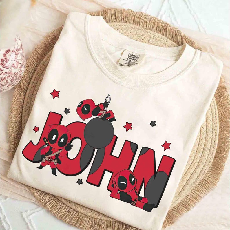 May include: Cream-coloured t-shirt with the name "JOHN" in red block letters and Deadpool-inspired illustrations. The design includes Deadpool characters and red and black stars. The shirt has a Comfort Colors tag.