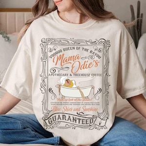 May include: Cream-colored t-shirt featuring a graphic with the text "VOODOO QUEEN OF THE BAYOU Mama Odie's APOTHECARY & TREEHOUSE COFFEE." The graphic includes an illustration of a character in a bathtub. The shirt also has the text "Blue Skies and Sunshine GUARANTEED."