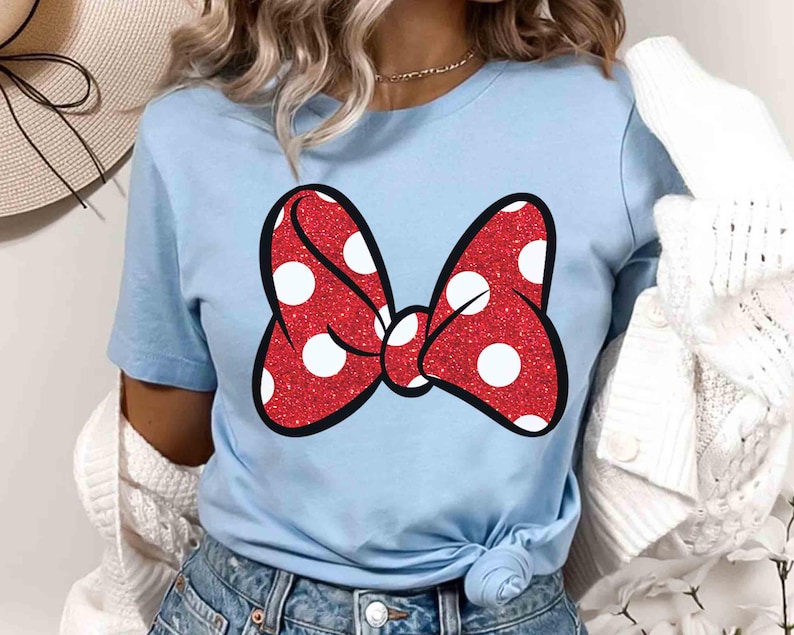 Puede incluir: Camiseta azul claro con un lazo rojo brillante con lunares blancos y contorno negro. El lazo est&aacute; centrado en la camiseta. La camiseta es de material suave y es adecuada para uso casual.