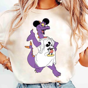 Könnte beinhalten: Cremefarbenes T-Shirt mit einem lila Drachen, der ein weißes Shirt mit einem Mickey Mouse-Motiv trägt. Der Drache hat schwarze Mauseohren und orangefarbene Akzente. Das Design ist eine verspielte Cartoon-Illustration.