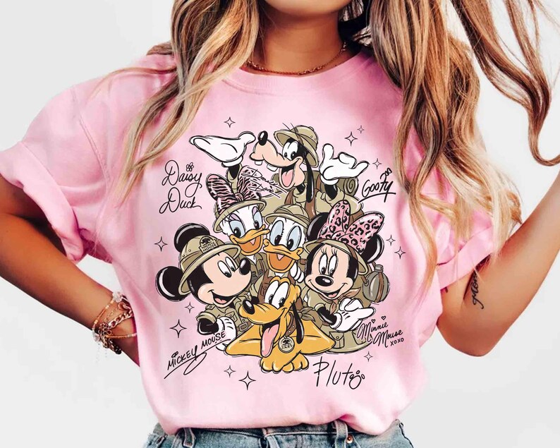May include: Pale pink t-shirt with a graphic of Disney characters Mickey Mouse, Minnie Mouse, Donald Duck, Daisy Duck, Goofy, and Pluto in safari outfits. The shirt includes character signatures and star accents.