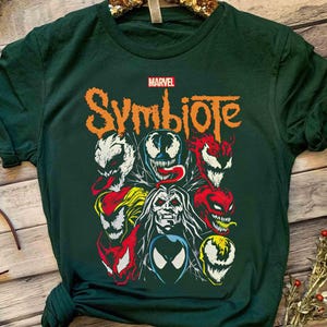 May include: Dark green t-shirt featuring a Marvel Symbiote design. The graphic includes various characters with vibrant colors like red, white, and yellow. The word "Symbiote" is in orange, with the Marvel logo above.