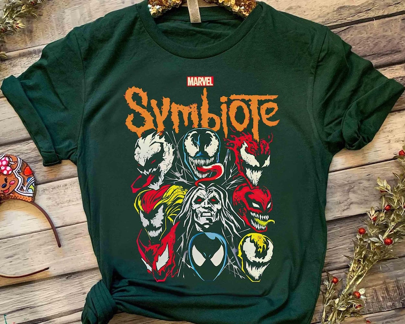 May include: Dark green t-shirt featuring a Marvel Symbiote design. The graphic includes various characters with vibrant colors like red, white, and yellow. The word "Symbiote" is in orange, with the Marvel logo above.