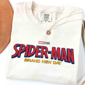 May include: Cream-colored t-shirt featuring the Marvel Studios Spider-Man logo in red and blue with yellow text that reads "BRAND NEW DAY." A gold necklace with a pendant is draped over the shirt.