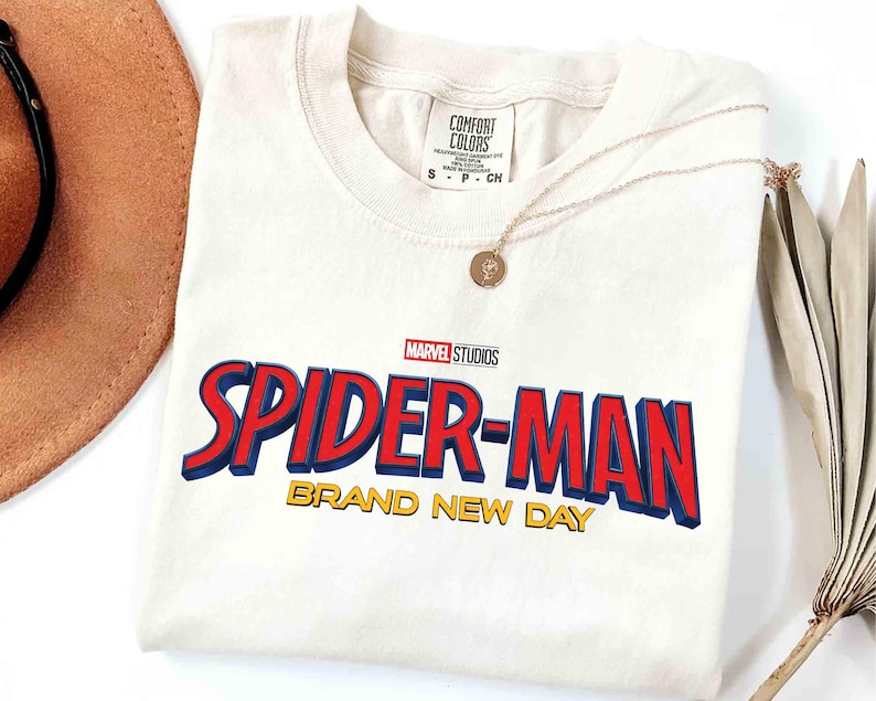 May include: Cream-colored t-shirt featuring the Marvel Studios Spider-Man logo in red and blue with yellow text that reads "BRAND NEW DAY." A gold necklace with a pendant is draped over the shirt.