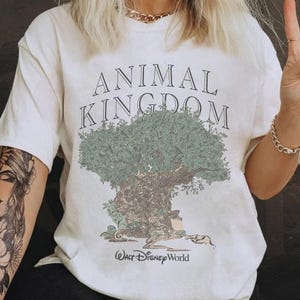 May include: A white short-sleeve t-shirt featuring a graphic of a large tree with green foliage and a brown trunk. The text ANIMAL KINGDOM is printed in black above the tree, and the text Walt Disney World is printed in black below the tree.