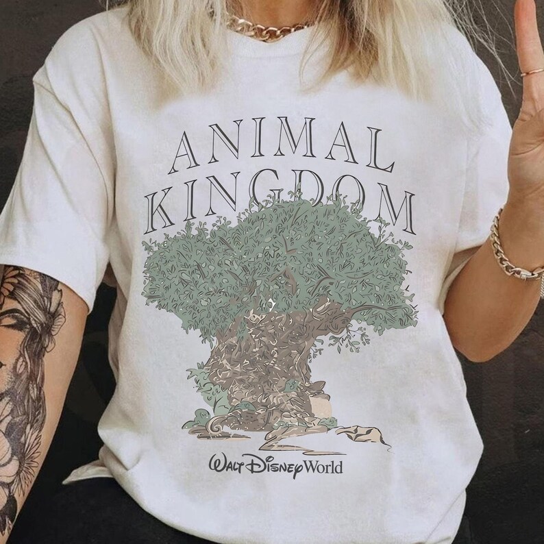 May include: A white short-sleeve t-shirt featuring a graphic of a large tree with green foliage and a brown trunk. The text ANIMAL KINGDOM is printed in black above the tree, and the text Walt Disney World is printed in black below the tree.