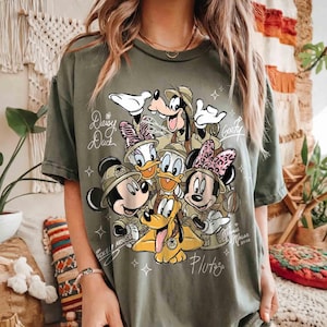 May include: An olive green t-shirt with a graphic of Disney characters dressed in safari outfits, including Mickey and Minnie Mouse, Donald and Daisy Duck, Goofy, and Pluto. The names of the characters are written on the shirt. Small star shapes surround the design.