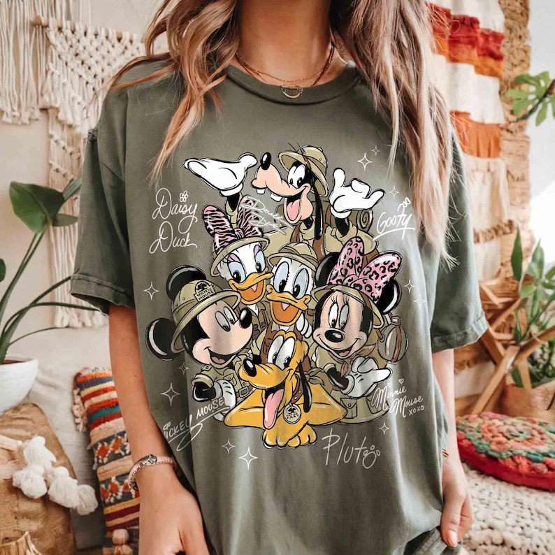 May include: An olive green t-shirt with a graphic of Disney characters dressed in safari outfits, including Mickey and Minnie Mouse, Donald and Daisy Duck, Goofy, and Pluto. The names of the characters are written on the shirt. Small star shapes surround the design.