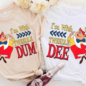 Könnte beinhalten: Zwei T-Shirts, eines in Beige und eines in Weiß, mit Cartoon-Illustrationen von Tweedledee und Tweedledum aus Alice im Wunderland. Die Texte "I'm With Tweedle Dum" und "I'm With Tweedle Dee" sind auf den Shirts aufgedruckt.