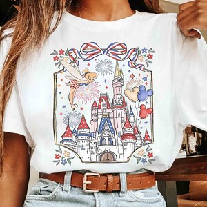 Puede incluir: Camiseta blanca con una colorida ilustraci&oacute;n de un hada, fuegos artificiales, un castillo y globos. El dise&ntilde;o incluye un lazo rojo, blanco y azul y detalles florales, creando un tema festivo y caprichoso.