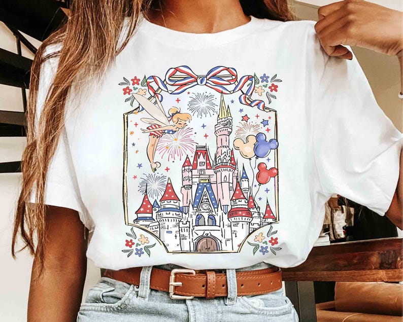 Puede incluir: Camiseta blanca con una colorida ilustraci&oacute;n de un hada, fuegos artificiales, un castillo y globos. El dise&ntilde;o incluye un lazo rojo, blanco y azul y detalles florales, creando un tema festivo y caprichoso.