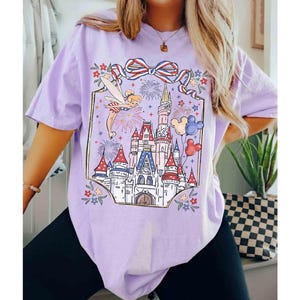 Puede incluir: Camiseta lila claro con un gr&aacute;fico colorido de un castillo de cuento de hadas, fuegos artificiales y un hada. El dise&ntilde;o incluye un lazo rojo, blanco y azul y globos de Mickey Mouse. La camiseta es holgada.