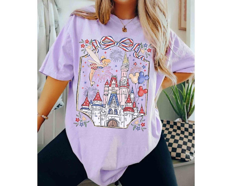 Puede incluir: Camiseta lila claro con un gr&aacute;fico colorido de un castillo de cuento de hadas, fuegos artificiales y un hada. El dise&ntilde;o incluye un lazo rojo, blanco y azul y globos de Mickey Mouse. La camiseta es holgada.