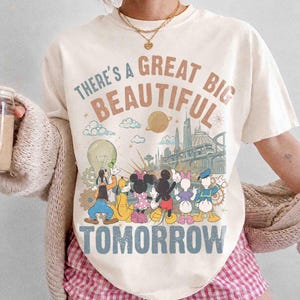 May include: An off-white short-sleeve t-shirt featuring a graphic print with the text "THERE'S A GREAT BIG BEAUTIFUL TOMORROW". The design includes illustrations of Goofy, Pluto, Minnie Mouse, Mickey Mouse, Daisy Duck, and Donald Duck.