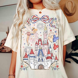 Puede incluir: Camiseta color crema con un gr&aacute;fico colorido de un hada, fuegos artificiales, un castillo y globos de Mickey Mouse. El dise&ntilde;o incluye un lazo rojo, blanco y azul y detalles florales, creando un aspecto festivo y caprichoso.