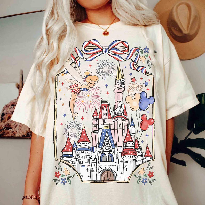 Puede incluir: Camiseta color crema con un gr&aacute;fico colorido de un hada, fuegos artificiales, un castillo y globos de Mickey Mouse. El dise&ntilde;o incluye un lazo rojo, blanco y azul y detalles florales, creando un aspecto festivo y caprichoso.