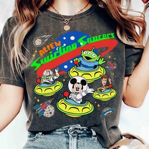 May include: Dark gray t-shirt featuring a colorful graphic of characters from the Toy Story franchise in flying saucers. The design includes Buzz Lightyear, Woody, Mickey Mouse, and the Little Green Men, with the text "Alien Swirling Saucers".