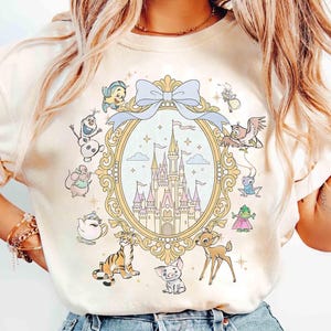 Puede incluir: Camiseta de color crema con un diseño caprichoso. Un castillo está en el centro, rodeado de personajes de dibujos animados. El diseño incluye un lazo azul claro y un marco dorado. Los personajes incluyen un tigre, un ciervo y otras figuras animadas.